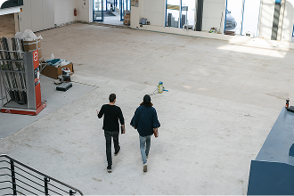 Zwei Personen gehen von oben fotografiert durch eine helle, leere Industriehalle oder einen Gewerbebau.