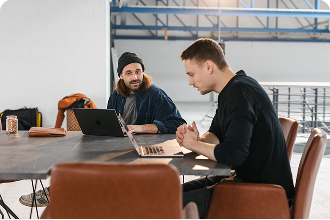 Zwei Männer sitzen an einem Tisch in einem Coworking-Space und arbeiten gemeinsam an einem Laptop.
